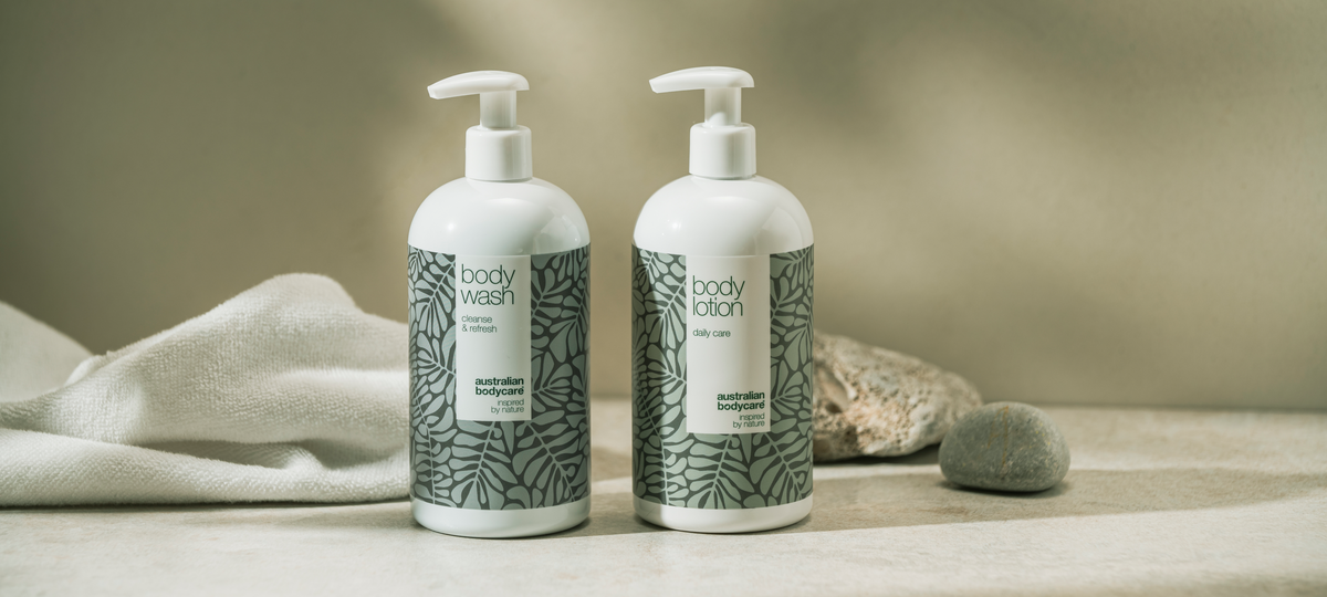 Body Wash a Body Lotion od Australian Bodycare ako veľké fľaše s pumpičkou v zelenom dizajne s tea tree, vedľa biely uterák a kamene.