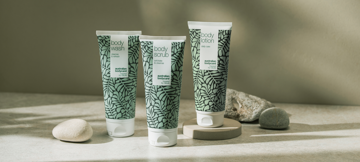 Body Wash, Body Scrub a Body Lotion od Australian Bodycare v zelenom dizajne s tea tree, postavené na svetlých kamenných podstavcoch na olivovom pozadí.