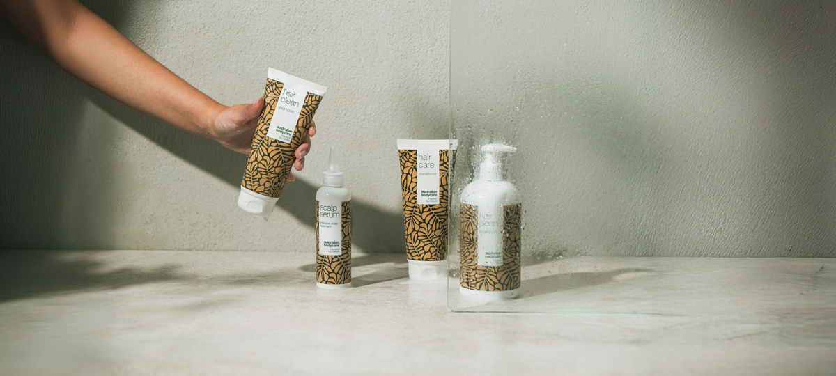 Ruka drží tubu Hair Clean Shampoo od Australian Bodycare pred sprchovou zástenou; pod ňou Scalp Serum, Hair Care Conditioner a fľaša Hair Clean s pumpičkou.