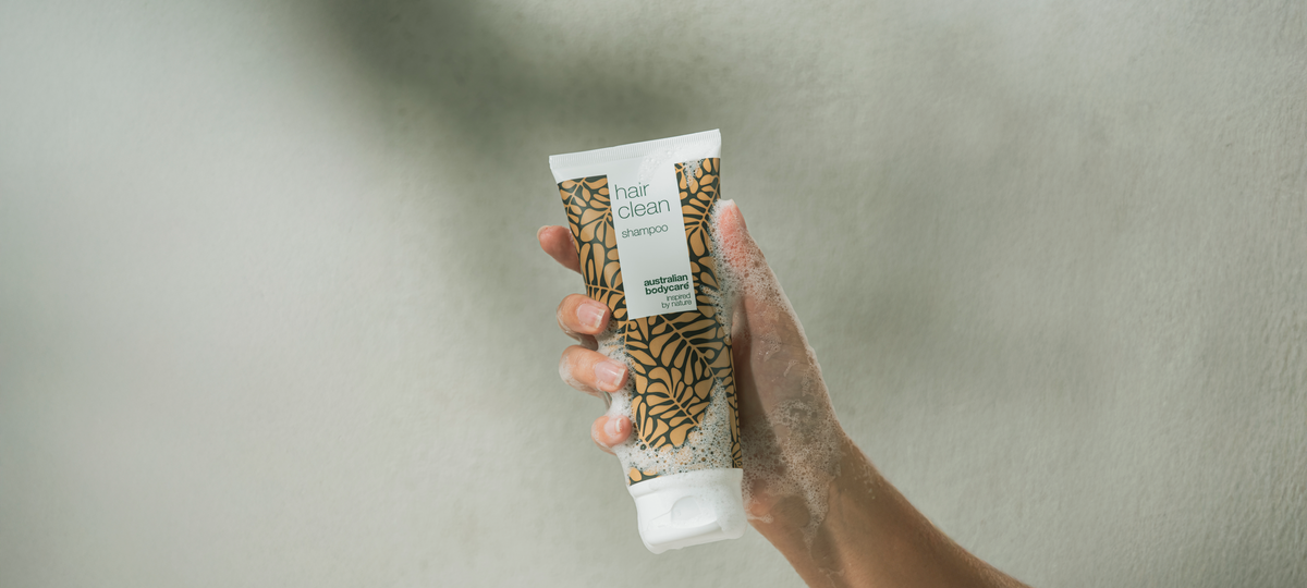 Zapenená ruka drží tubu Hair Clean Shampoo od Australian Bodycare v béžovom obale v sprche.
