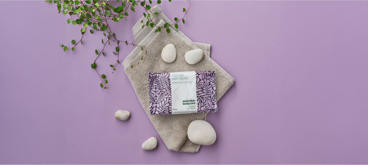 Balenie Intimate Wet Wipes od Australian Bodycare na béžovom uteráku na fialovom pozadí, okolo biele okruhliaky a zelená vetvička.