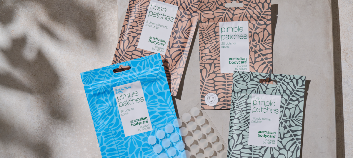 Niekoľko balení náplastí od Australian Bodycare cez seba na kamennom povrchu: Nose Patches a Pimple Patches v béžovej, Salicylic Acid Pimple Patches v modrej a Body Pimple Patches v zelenej, vedľa hárok s náplasťami.