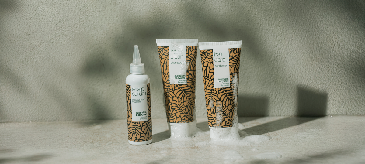 Scalp Serum, Hair Clean Shampoo a Hair Care Conditioner od Australian Bodycare v béžovom obale na kamennom povrchu, okolo túb pena.
