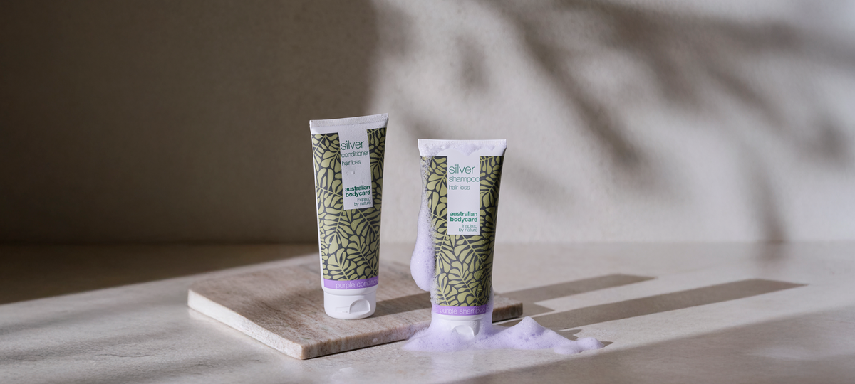 Silver Conditioner a Silver Shampoo od Australian Bodycare s fialovým lemom na mramorovej doske, vedľa fialovo sa lesknúcej peny.